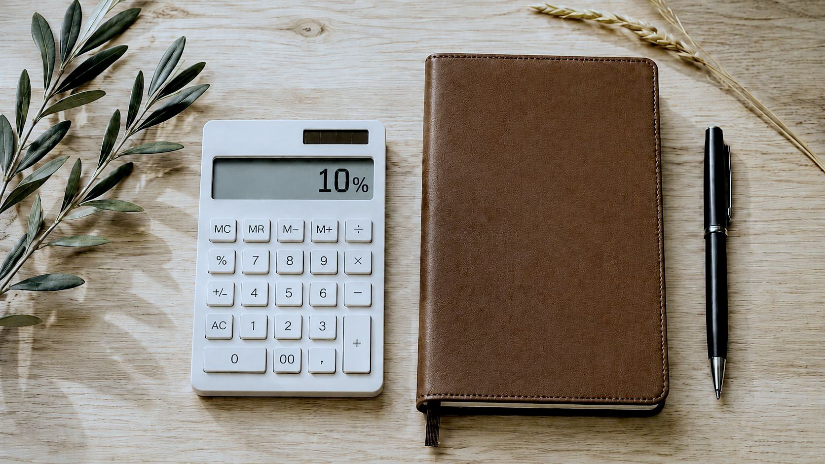 Calculadora mostrando 10% ao lado de um caderno de anotações e um ramo de oliveira, sobre mesa de madeira clara