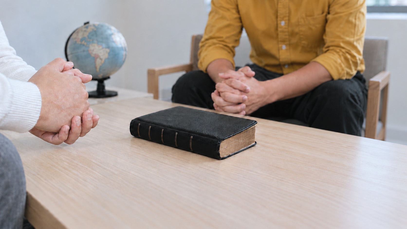 Duas pessoas com as mãos postas em oração à mesa, com uma Bíblia e um globo terrestre ao lado, em culto de missões